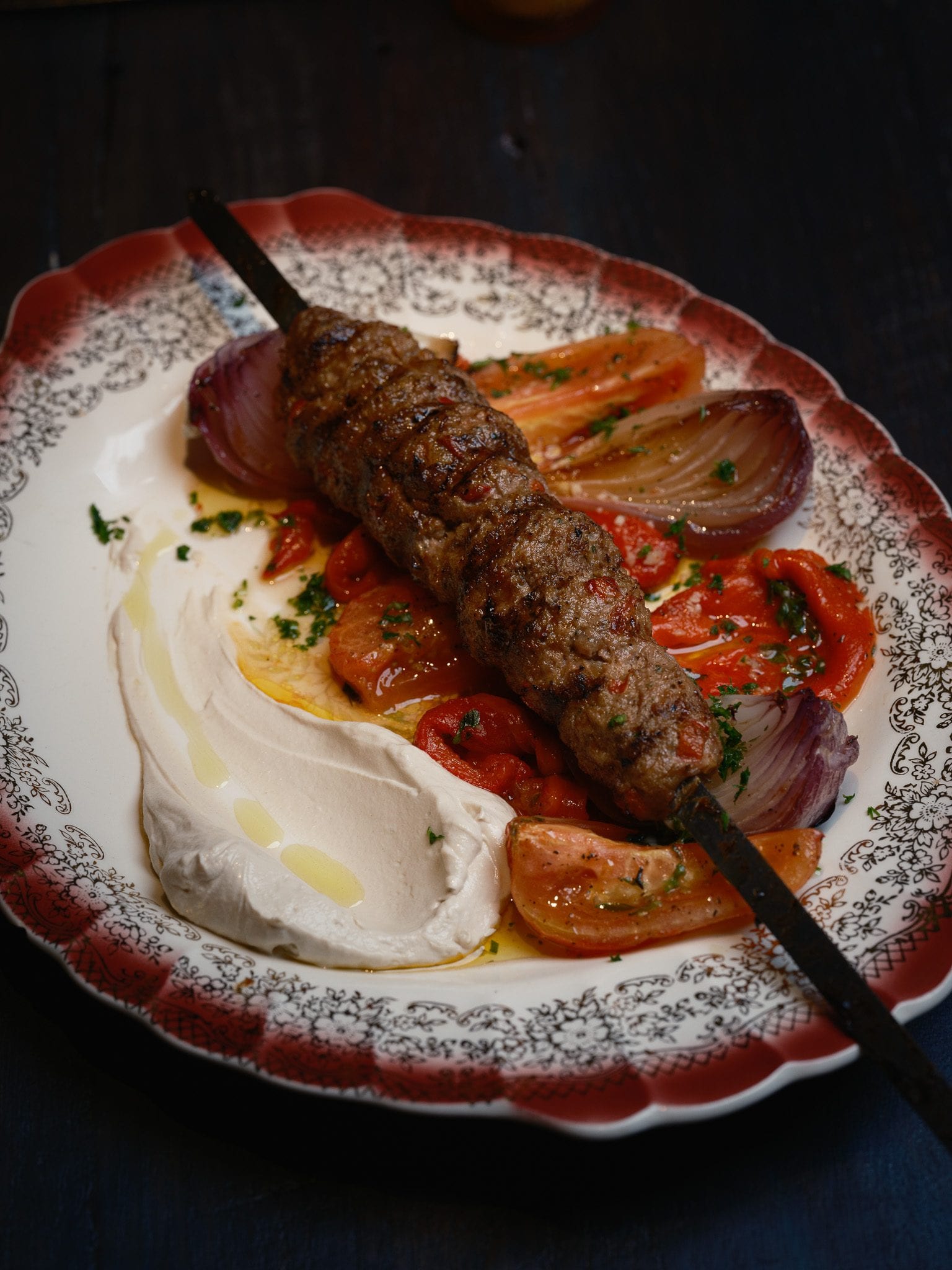 Lamb lula kebab at GUSI over labneh with roasted peppers, tomato, and onion on a patterned platter